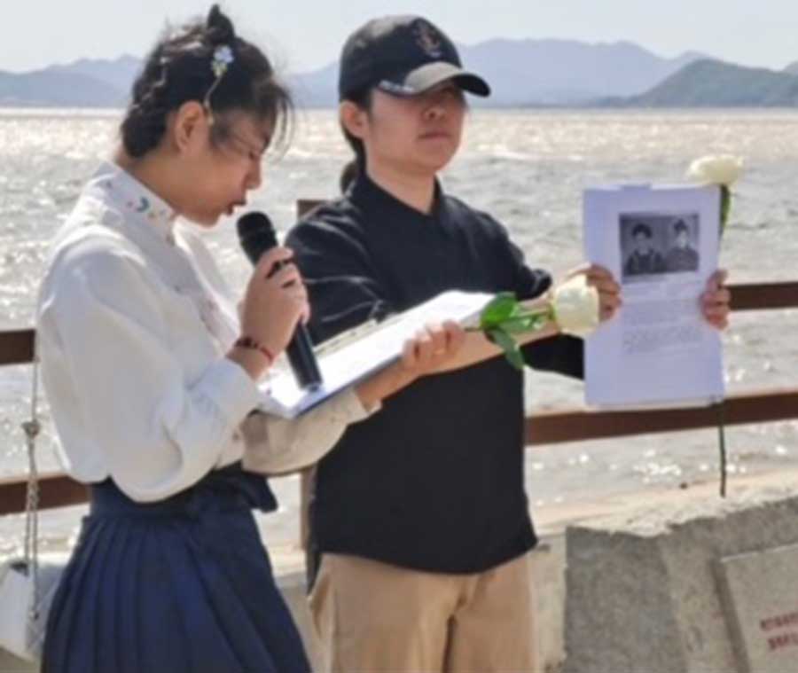 A young local girl exhibits a photograph while another narrates a tribute written by Anne Kelly to her Uncle Maurice Kelly, 2nd of October 2022, the 80th anniversary of the sinking of the Lisbon Maru, at the site of the memorial for the rescue of British Prisoners of War by Dongji fishermen.